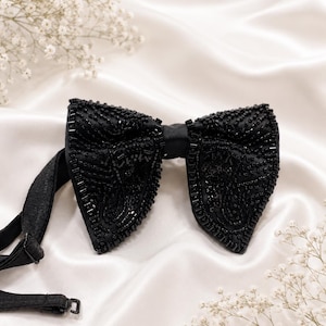 May include: A black beaded bow tie with a satin finish. The bow tie is embellished with small, dark beads and has an adjustable strap. The background is a white satin fabric with small white flowers.