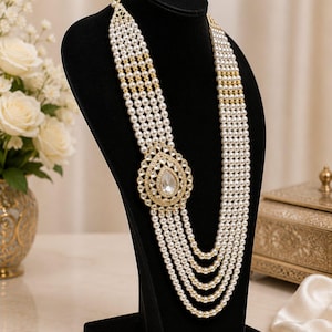 May include: A multi-strand necklace featuring five rows of white pearls accented with small gold-toned beads. The necklace includes a large, oval-shaped gold-toned pendant set with clear stones and a central teardrop-shaped stone at the center.