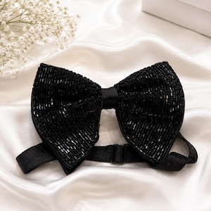 May include: A black beaded bow tie with an adjustable strap. The bow tie is made of small, shiny beads and is set against a white satin background. A formal accessory.