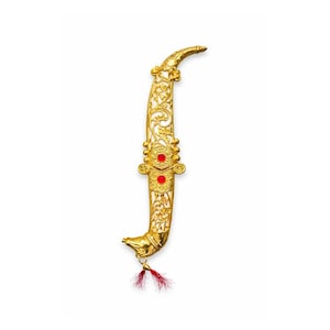 May include: A gold-colored, ornate dagger with intricate floral patterns and two red gemstone accents. The curved blade features a decorative design, and a small tassel hangs from the hilt. The dagger is set against a white background.