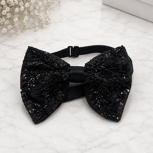 May include: A black sequined bow tie with a satin centre. The bow tie is on a black adjustable strap. The background is a marble surface with white flowers and a white box.