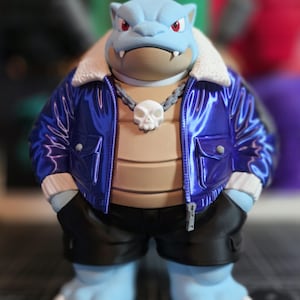 May include: A light blue creature figurine with red eyes and white claws, wearing a shiny metallic blue bomber jacket with a white faux-fur collar, a white skull pendant on a grey chain, and black shorts, standing in a forward-facing pose.