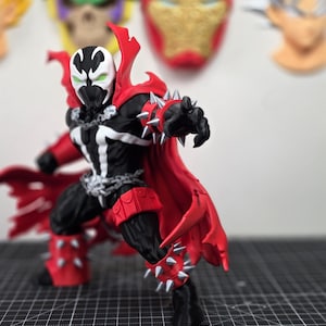 May include: A Spawn action figure featuring a black and white suit with a flowing red cape, silver spiked gauntlets, and silver spiked boots. The character has a white mask with green eyes and is adorned with silver chains around the chest and waist.