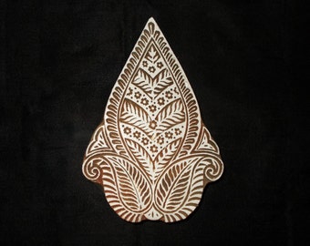 May include: A wooden block printing stamp with a white intricate design of leaves and flowers. The stamp is brown and has a teardrop shape.