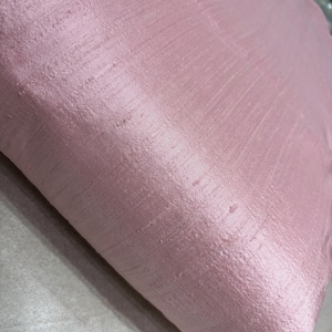 May include: A close-up shot of a dusty rose-colored fabric with a subtle sheen. The material has a textured surface with visible threads and slight imperfections, suggesting a natural textile. The fabric is folded, creating shadows and highlighting its depth.