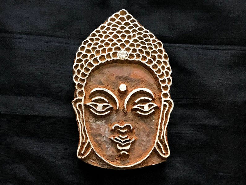 May include: A wooden block carving of a Buddha's face with a traditional design. The carving is detailed and intricate, with a serene expression.