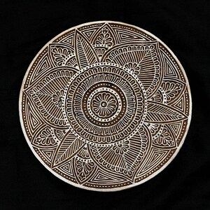 May include: A white circular wooden block with a detailed mandala design carved into the surface. The mandala features intricate patterns of leaves, flowers, and geometric shapes.