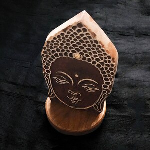May include: A wooden sculpture of a Buddha's face with a brown and black design. The sculpture is on a wooden base.