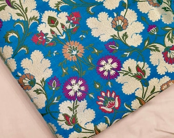 Könnte beinhalten: Nahaufnahme eines blauen Stoffes mit einem weißen und goldenen Blumenmuster. Der Stoff hat eine Brokat-Textur und weist rosa, rote und violette Akzente auf.