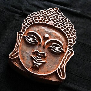 May include: A wooden block carving of a Buddha's face with intricate details. The carving is brown and has a raised design.