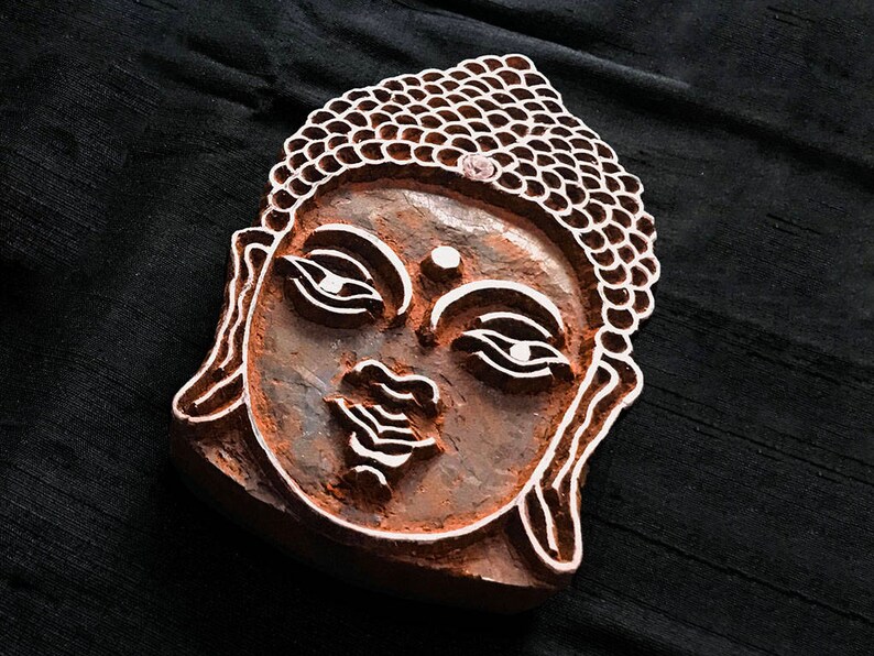 May include: A wooden block carving of a Buddha's face with intricate details. The carving is brown and has a raised design.
