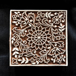 Puede incluir: Un bloque de impresión de madera con un diseño floral detallado. El diseño presenta una gran flor central rodeada de flores y hojas más pequeñas. El bloque está tallado en un color marrón claro y tiene un fondo blanco.