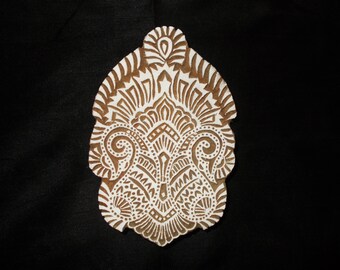 May include: A white wooden block with a detailed, intricate design carved into it. The design is a symmetrical pattern of swirls and leaves, reminiscent of a paisley pattern.