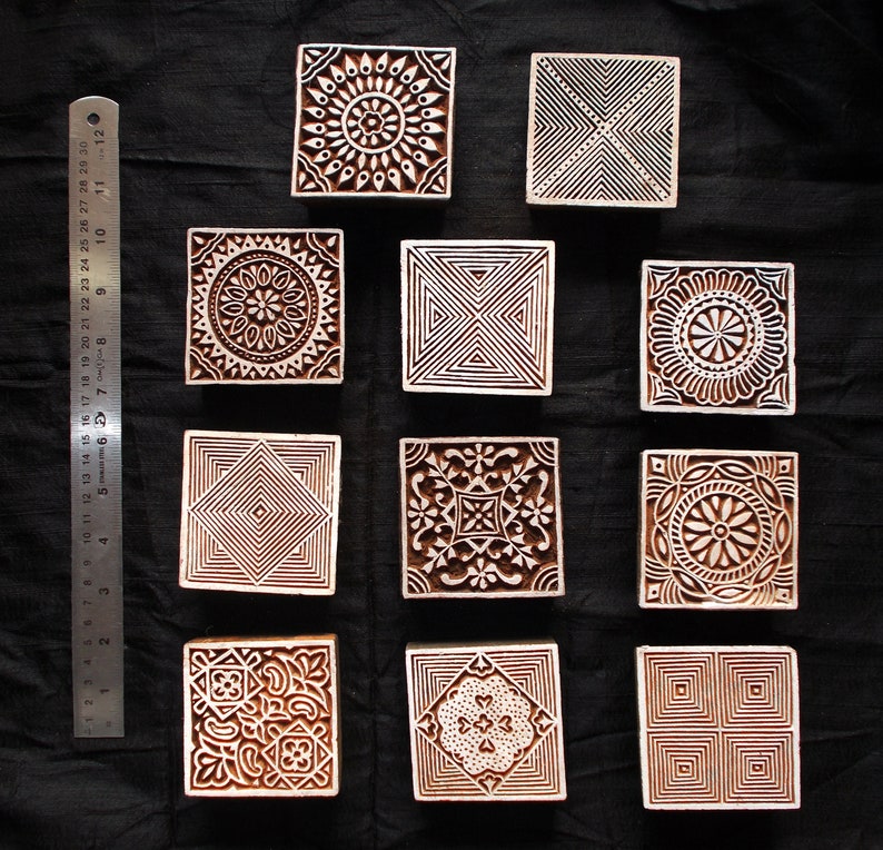 Puede incluir: Una colecci&oacute;n de once bloques de impresi&oacute;n cuadrados de madera con intrincados dise&ntilde;os tallados. Los bloques presentan patrones geom&eacute;tricos y florales en tonos marrones. Una regla con medidas en cent&iacute;metros es visible a la izquierda.
