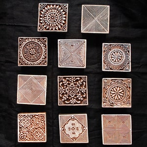 Puede incluir: Una colección de once bloques de impresión cuadrados de madera con intrincados diseños tallados. Los bloques presentan patrones geométricos y florales en tonos marrón y blanco, dispuestos sobre un fondo oscuro.