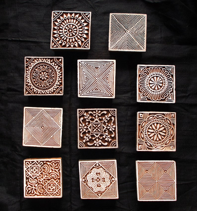 Puede incluir: Una colecci&oacute;n de once bloques de impresi&oacute;n cuadrados de madera con intrincados dise&ntilde;os tallados. Los bloques presentan patrones geom&eacute;tricos y florales en tonos marr&oacute;n y blanco, dispuestos sobre un fondo oscuro.