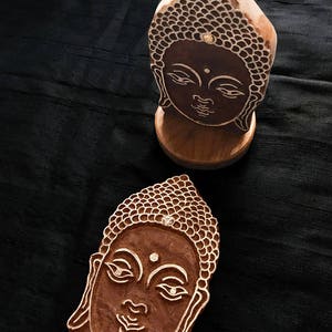 May include: Two wooden Buddha head carvings, one standing on a base and the other lying flat. Both carvings feature intricate details and a serene expression.