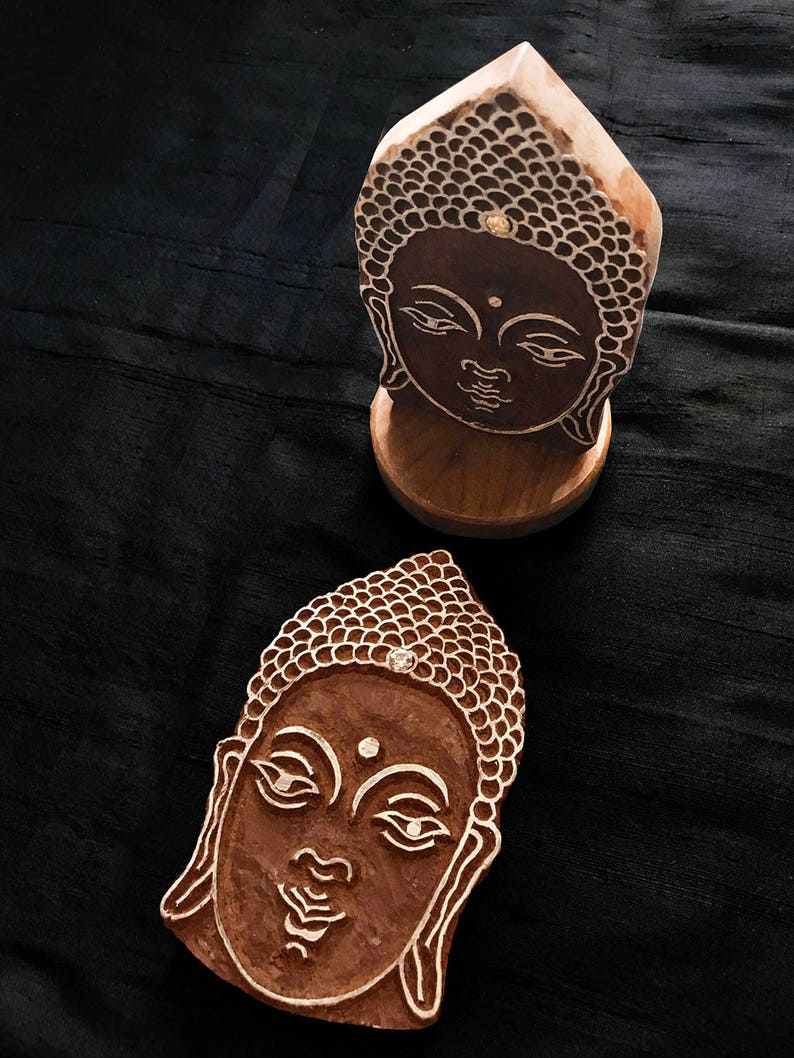 May include: Two wooden Buddha head carvings, one standing on a base and the other lying flat. Both carvings feature intricate details and a serene expression.