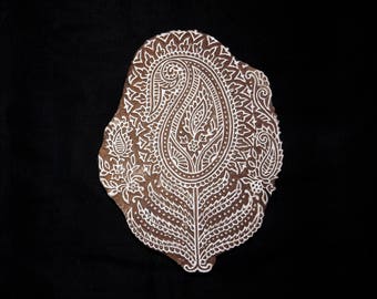 May include: A white paisley design carved into a brown wooden block. The design is intricate and detailed, with a central motif of a large paisley shape surrounded by smaller paisley shapes and floral elements.