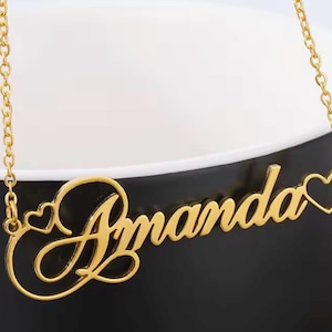 Può includere: Una collana con targhetta dorata che riporta il nome Amanda in carattere corsivo. Il design include il contorno di un piccolo cuore sul lato sinistro e il contorno di un cuore più grande sul lato destro, collegati a una sottile catenina dorata.