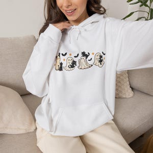 Può includere: Una felpa con cappuccio bianca caratterizzata da una stampa grafica di quattro fantasmi in stile cartoon e diversi gatti neri. Il design include piccole stelle arancioni e pipistrelli neri sparsi tra le figure. La felpa ha maniche lunghe.