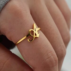 Può includere: Un anello color oro caratterizzato da una lettera A stilizzata e prominente in carattere corsivo. La fascia è sottile e liscia, collegata alla base della lettera. Il design è lucido e riflettente, con una finitura in oro giallo brillante.