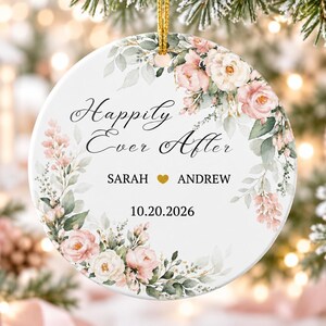 Peut inclure: Une décoration ronde en céramique blanche ornée d'une couronne florale de roses roses et blanches avec des feuilles vertes. Le texte indique Happily Ever After, Sarah, une icône de cœur doré, Andrew et la date 10.20.2026. Elle est suspendue par un cordon doré.