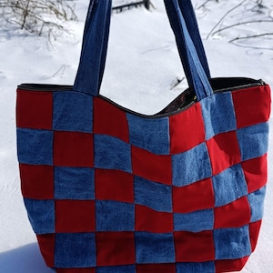 Puede incluir: Un bolso tote de patchwork con un patrón de tablero de ajedrez de cuadrados de mezclilla rojos y azules. El bolso tiene asas de mezclilla azules y un cierre de cremallera negro. El bolso está sobre una superficie nevada.