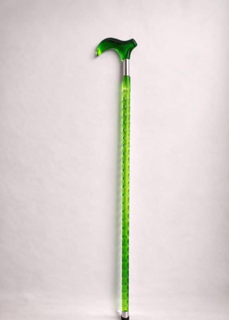 May include: A green walking cane with a curved handle and a textured shaft. The handle and the top of the shaft are a darker shade of green, while the shaft has a lighter green color. The cane has a silver metal band and a silver tip.