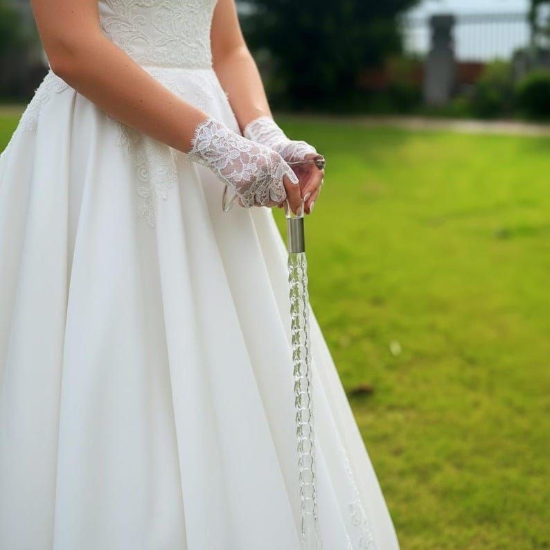 May include: A bride in a white wedding dress and lace gloves holds a clear, decorative wand. The wand is made of clear, faceted crystals and has a silver handle. The background is a blurred green lawn.