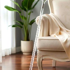May include: A clear, decorative walking cane with a silver-colored handle rests against a cream-colored armchair. The cane's shaft has a textured, spiral design. A potted plant is in the background.