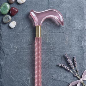 May include: A pink, decorative cane with a curved handle and a textured shaft. The cane has a gold-colored band at the top. The image also includes decorative stones and lavender sprigs.