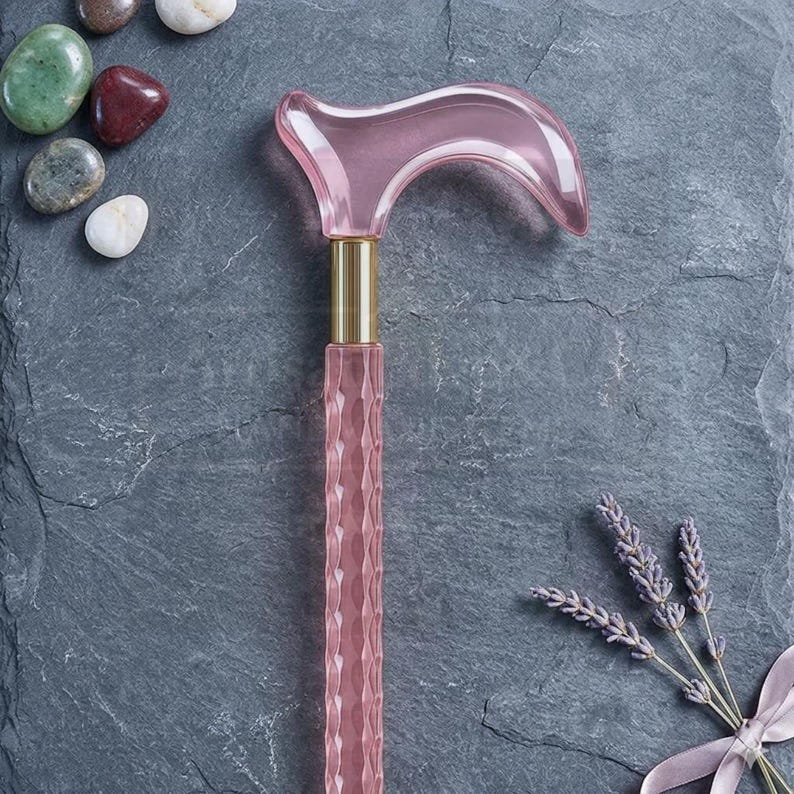 May include: A pink, decorative cane with a curved handle and a textured shaft. The cane has a gold-colored band at the top. The image also includes decorative stones and lavender sprigs.