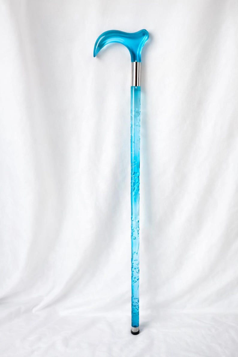 May include: A transparent blue walking cane with a curved handle. The handle and shaft are a vibrant blue color, with a silver metal band connecting the handle to the shaft. The shaft has a decorative pattern.