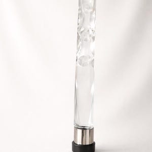 May include: A clear, textured glass bottle stopper with a silver metal base and a black rubber bottom. The glass has a unique, irregular shape with rounded indentations. The stopper is designed to fit into a bottle.