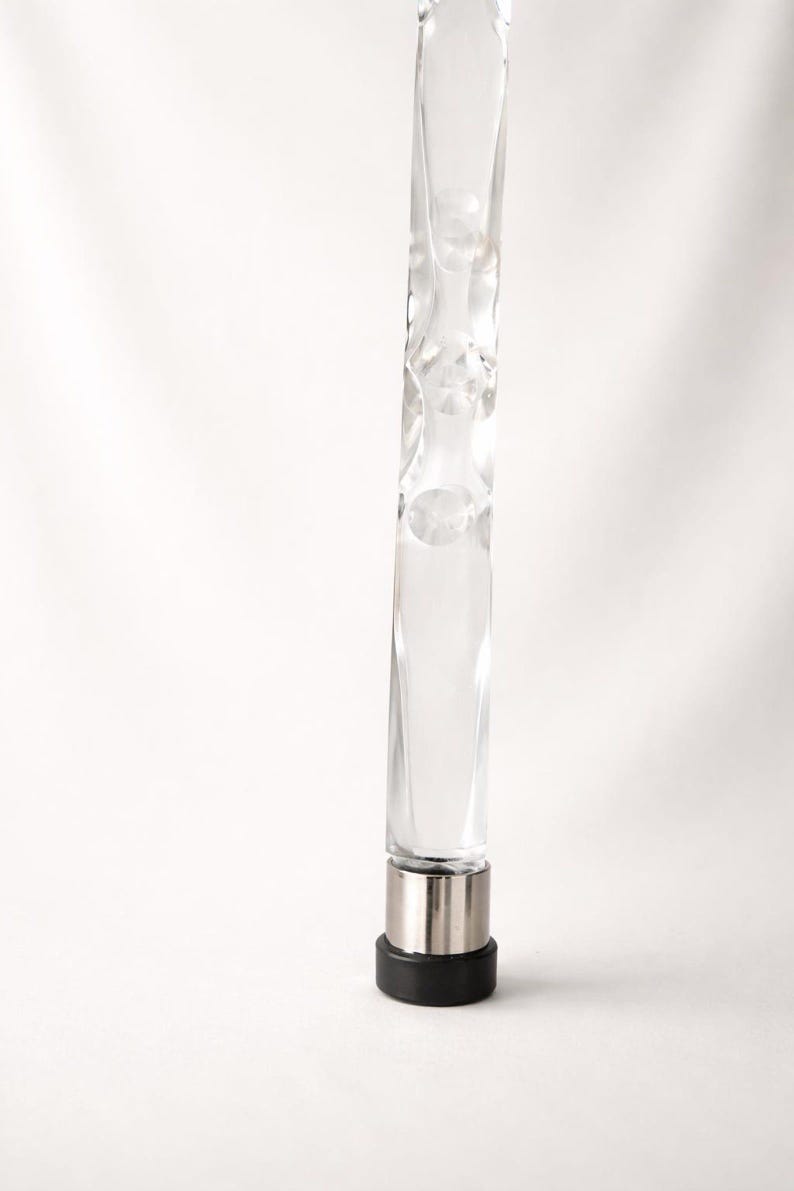 May include: A clear, textured glass bottle stopper with a silver metal base and a black rubber bottom. The glass has a unique, irregular shape with rounded indentations. The stopper is designed to fit into a bottle.