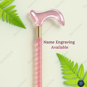 May include: A pink walking cane with a curved handle and a textured shaft. The handle is a transparent pink, and the shaft has a diamond pattern. The text "Name Engraving Available" is visible.