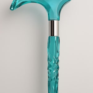 May include: A turquoise walking cane with a curved handle and a textured shaft. The handle is a solid, smooth shape, while the shaft has a rippled, organic design. A silver-colored metal band connects the handle and shaft.