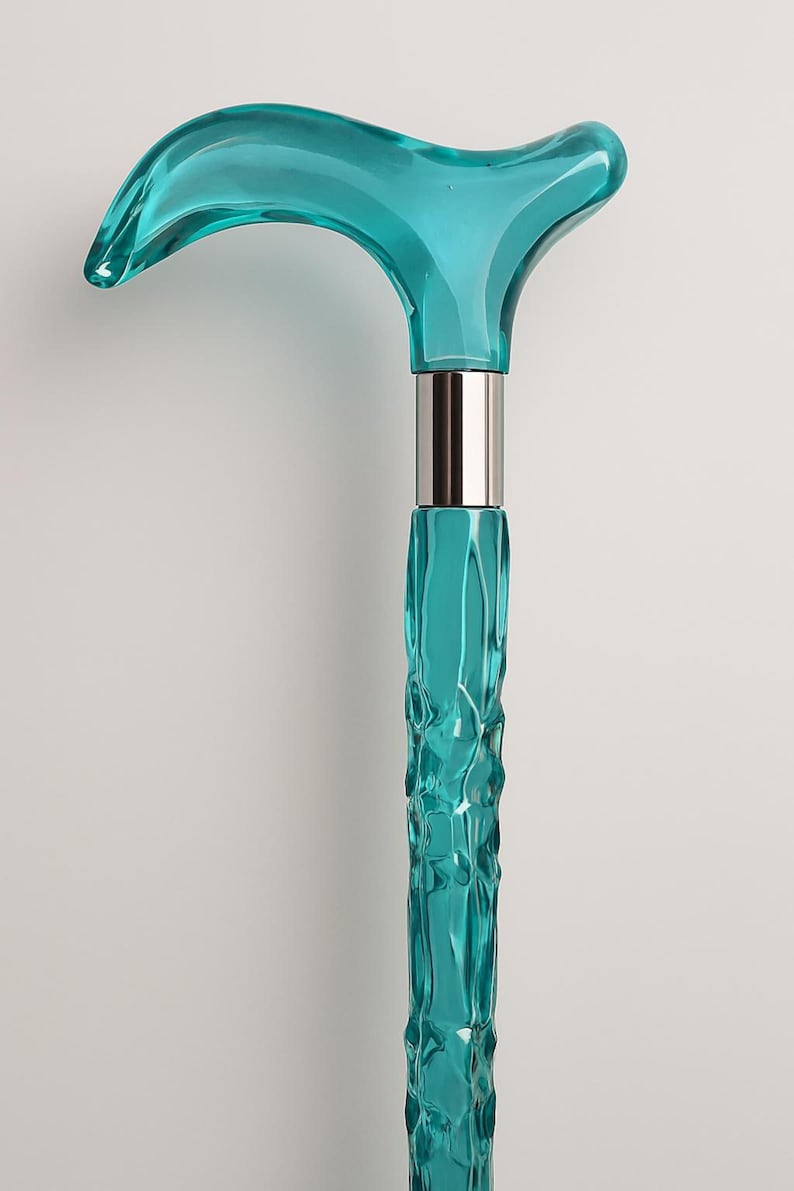 May include: A turquoise walking cane with a curved handle and a textured shaft. The handle is a solid, smooth shape, while the shaft has a rippled, organic design. A silver-colored metal band connects the handle and shaft.