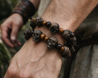 Pode incluir: Duas pulseiras elásticas com contas de caveira esculpidas em castanho escuro alternadas com contas redondas lisas e polidas na cor âmbar. As contas de caveira apresentam detalhes faciais precisos, incluindo órbitas oculares e dentes.