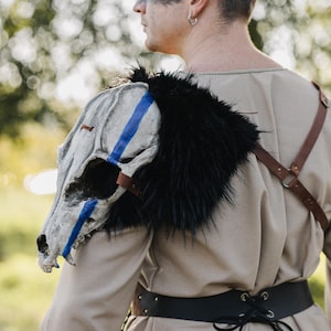 May include: A shoulder piece featuring a painted animal skull with blue stripes, black fur trim, and brown leather straps. The skull is white and gray, and the fur is black. The person is wearing a tan tunic and a black belt.