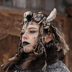 May include: A headpiece constructed from animal bones, feathers, and wooden beads, worn with a dark brown faux fur collar and a grey textured shawl. The accessory features a central skull element and dangling strands of beads framing the face.