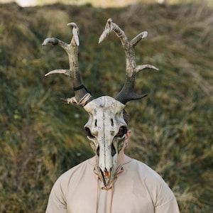 May include: A detailed, handcrafted mask resembling a deer skull with large antlers. The mask is primarily off-white with dark accents and features intricate markings. The antlers are a mix of brown and grey, and the mask is worn by a person.