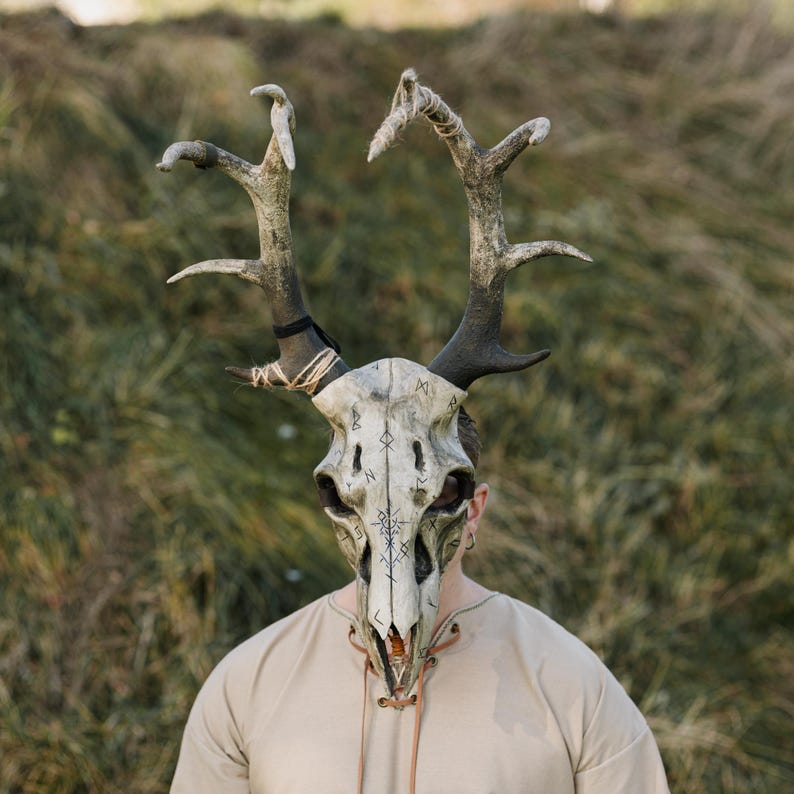 May include: A detailed, handcrafted mask resembling a deer skull with large antlers. The mask is primarily off-white with dark accents and features intricate markings. The antlers are a mix of brown and grey, and the mask is worn by a person.