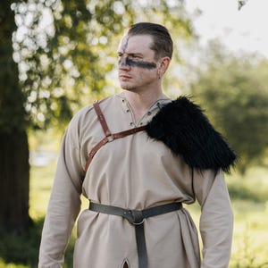 May include: A beige long-sleeved tunic with a lace-up neckline, paired with a dark brown leather chest harness featuring metal rings. A black faux fur shoulder mantle is attached to the harness, and a dark brown leather belt is worn at the waist.