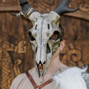 May include: A detailed skull mask with dark antlers and rune-like markings. The mask is painted in shades of grey and off-white, with dark blue symbols. The antlers are black and attached with twine. A leather strap secures the mask.