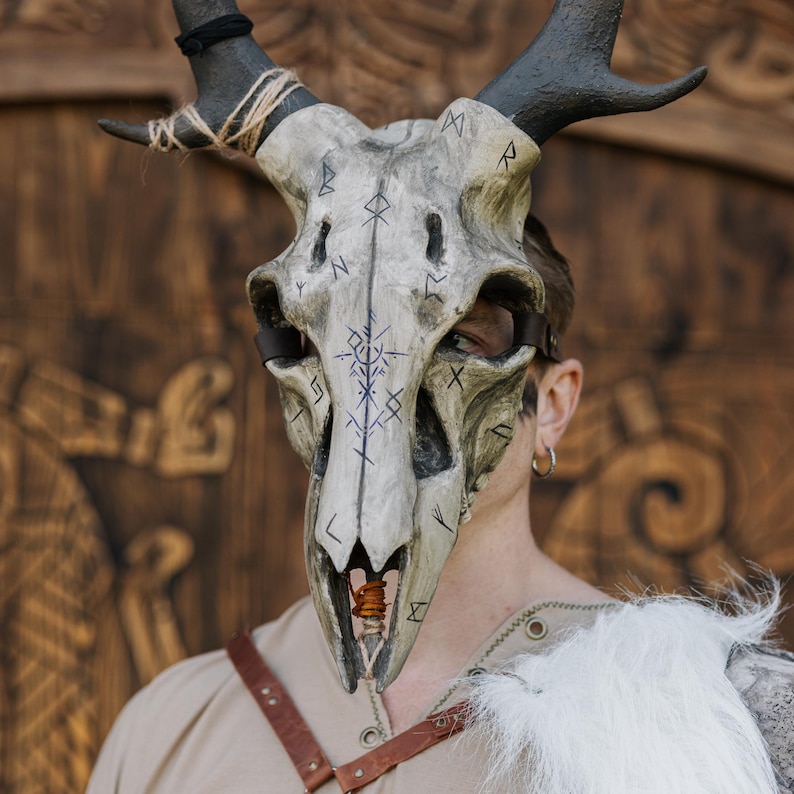 May include: A detailed skull mask with dark antlers and rune-like markings. The mask is painted in shades of grey and off-white, with dark blue symbols. The antlers are black and attached with twine. A leather strap secures the mask.