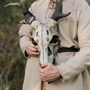 May include: A detailed, weathered skull mask with large, dark antlers. The skull is painted with runes and symbols, and is held by a person wearing a tan tunic. The antlers are wrapped with twine and secured with black straps.