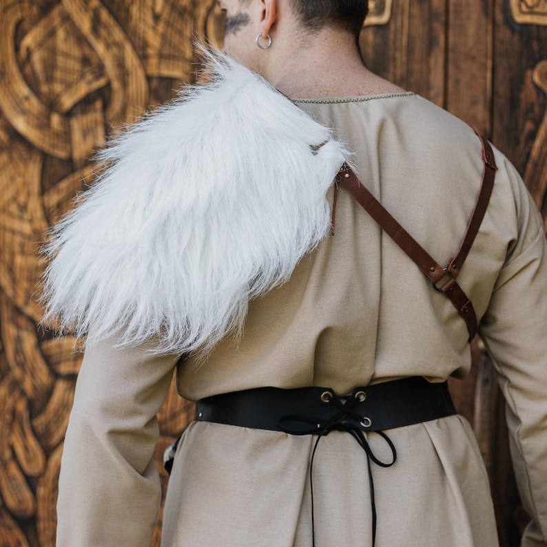 May include: A beige long-sleeved tunic worn with a white faux fur shoulder mantle, a brown leather strap harness across the chest, and a wide black leather waist belt featuring a lace-up front closure with black cord ties.