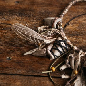 May include: A necklace featuring a braided cord adorned with brown and white patterned feathers, light-colored tooth-shaped pendants, small wooden beads, and thin leather cords. Some of the tooth-shaped pendants are wrapped in black thread.